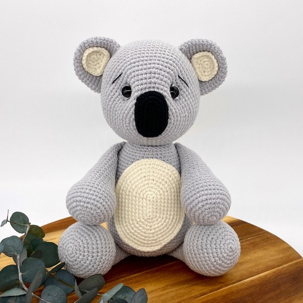 An amigurumi koala toy sitting on a wooden board with gum leaves.