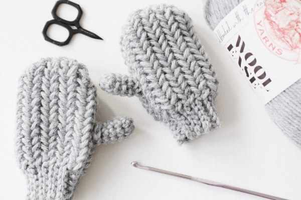 A flat lay image of a pair of light grey childs crochet mittens with crochet supplies.