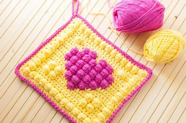 A bright yellow crochet potholder with a fuschia pink heart motif.