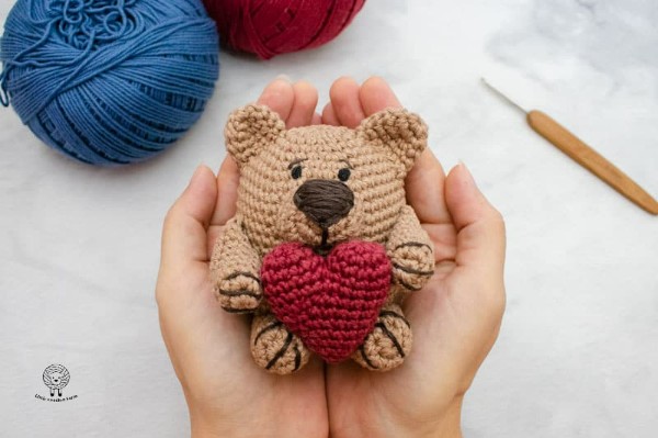 A small brown crocheted teddy bear holding a red heart.