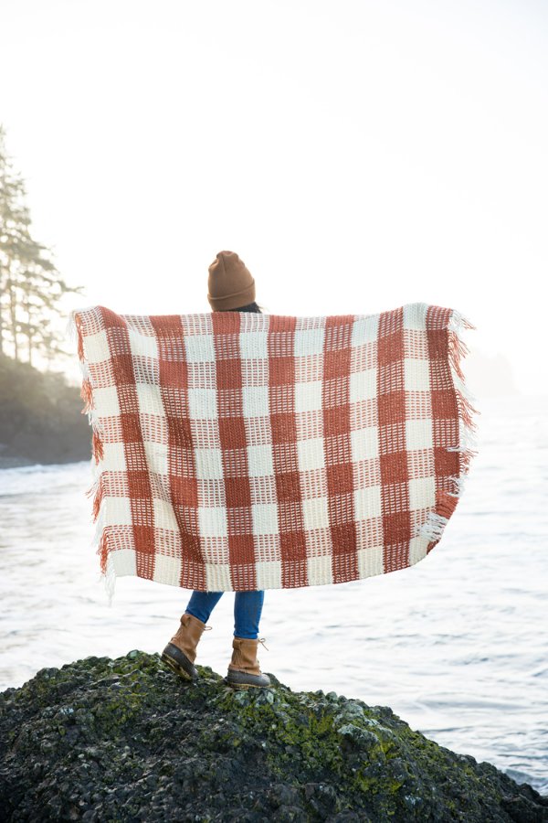 A woman standing by the ocean while holding out a crochet gingham blanket.
