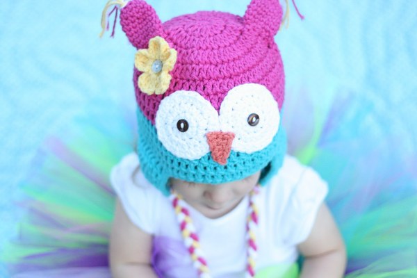 A young child wearing a brightly coloured crochet owl hat with earflaps and braided ties.