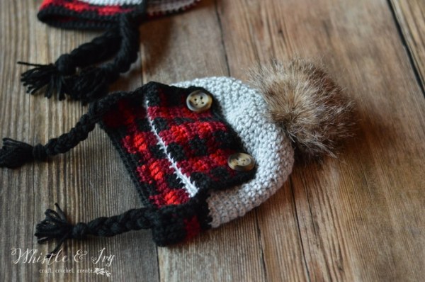 A crochet trapper-style baby hat with a plaid design and faux-fur pompom.