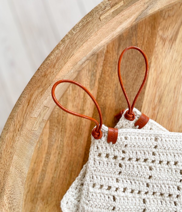 Two filet crochet potholders with leather loops on a timber board.