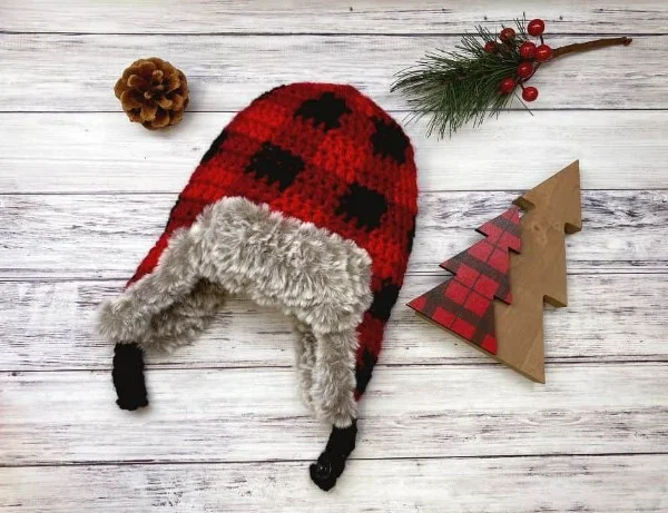 A flat lay image of a plaid colourwork crochet baby hat with earflaps.