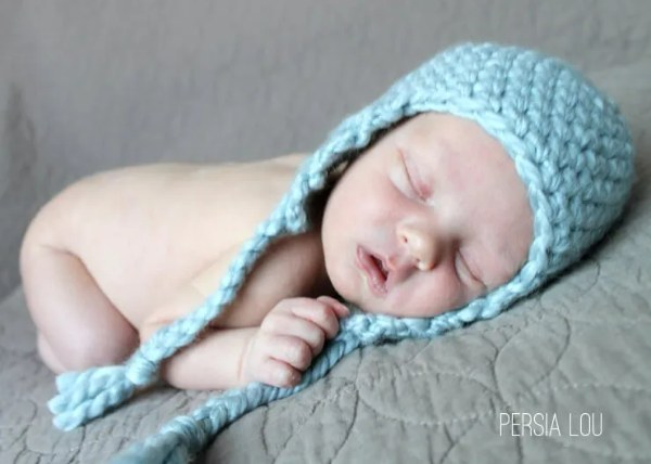A newborn baby wearing a blue crochet hat with earflaps.