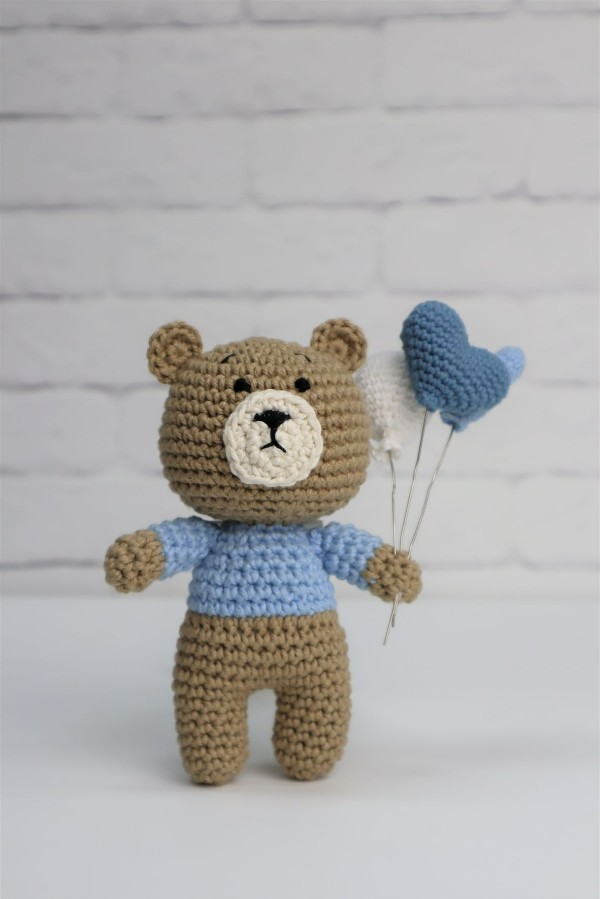 A brown crochet bear with a blue shirt and a bunch of heart-shaped crochet balloons.