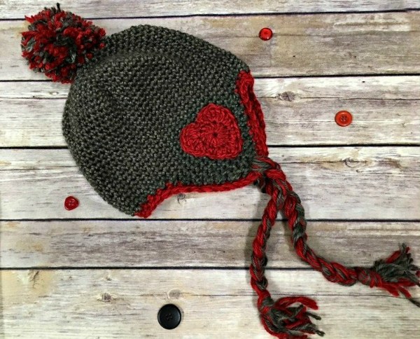 A flat-lay image of a grey crochet earflap hat with red trim and a heart applique.
