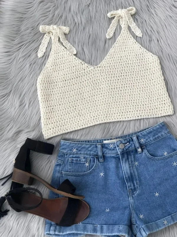 A flat lay image of a white crochet v-neck top with tie staps, jeans, and sandals.