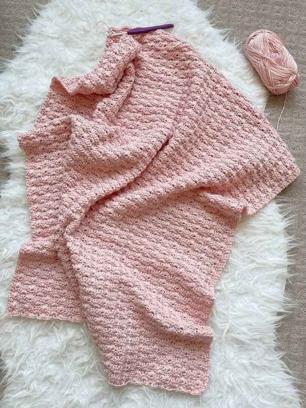 A pink shell stitch baby blanket on a white fur rug.