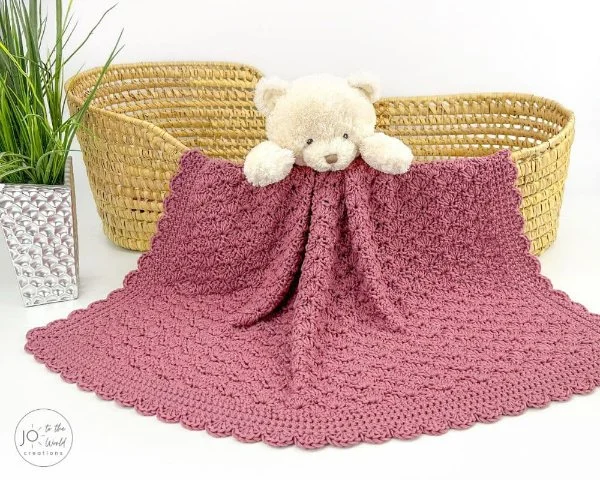 A moses basket with a teddy bear and a pink crocheted shell stitch baby blanket.