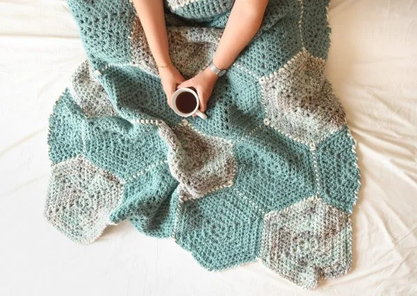 A closeup image of a blue and grey crochet hexagon blanket draped over a persons legs.