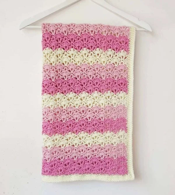 A pink and white striped shell stitch baby blanket on a wooden hanger.