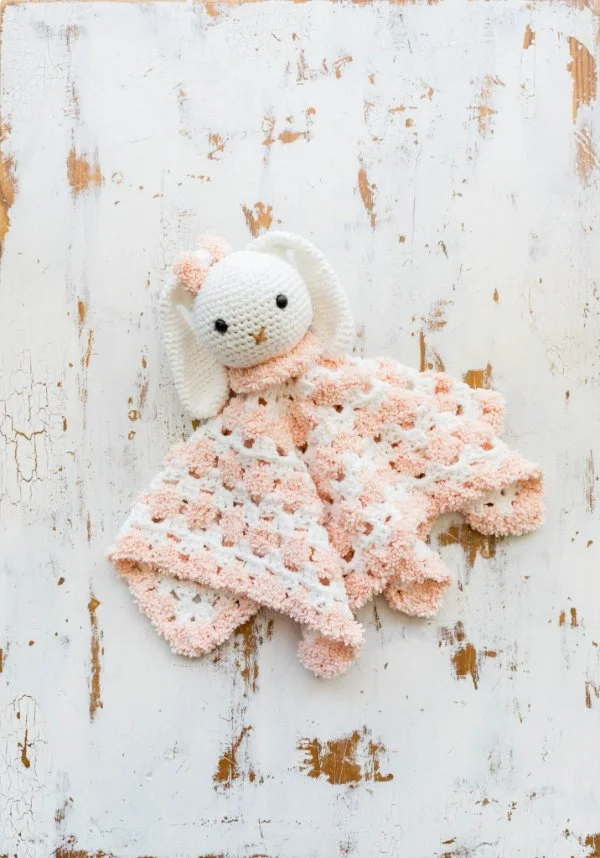 A pink and white crocheted bunny lovey on a white timber board background.