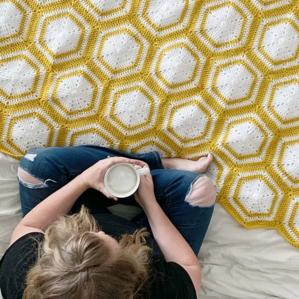 A person sitting on a bed with a hot drink, next to a yellow and white crochet hexagon blanket.
