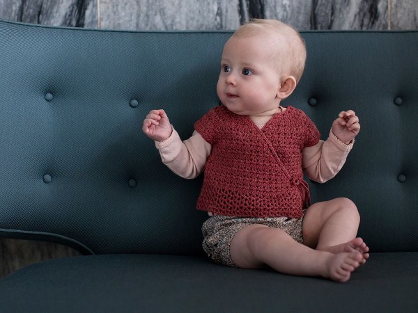 A baby wearing a red crochet wrap top and sitting on a sofa.