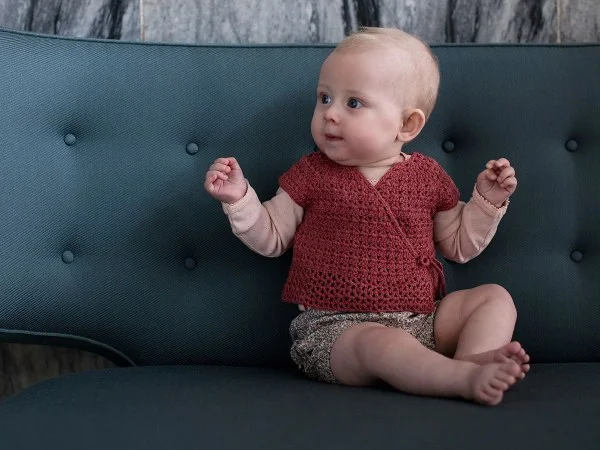 A baby wearing a red crochet wrap top and sitting on a sofa.