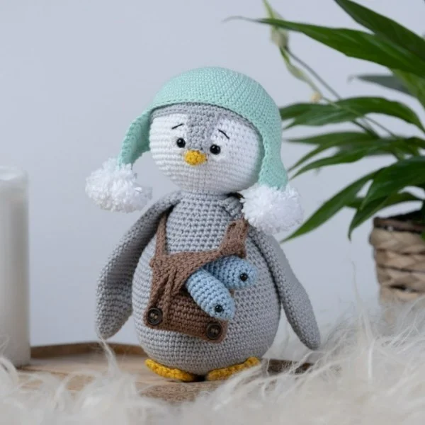 A crochet penguin earing an earflap hat and a bag with fish.