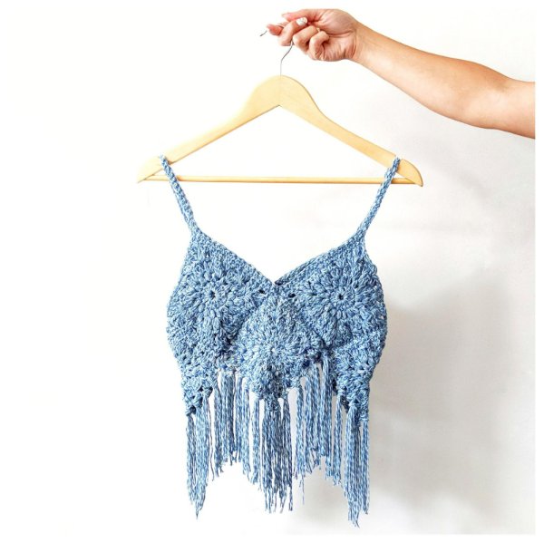 A blue granny square top with fringing hanging from a wooden hanger.