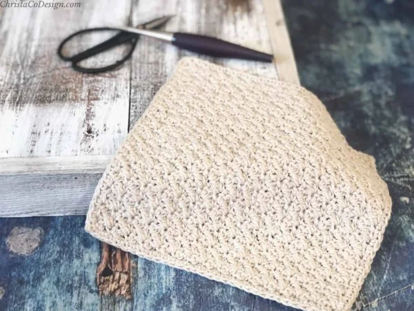 A cream coloured washcloth next to a crochet hook and black scissors.