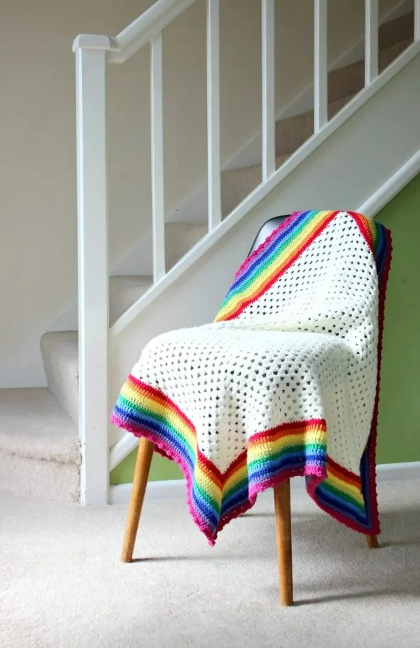 A white granny square blanket with a rainbow-striped border.