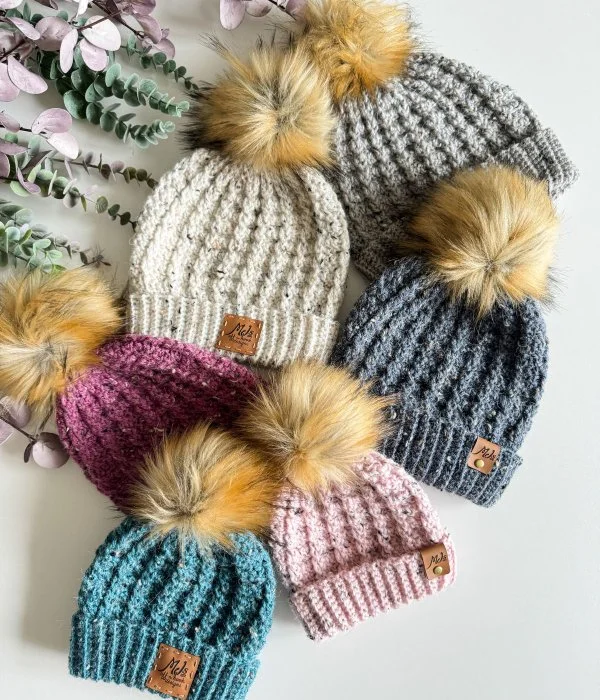 A pile of different coloured cable crochet hats with faux fur pompoms.