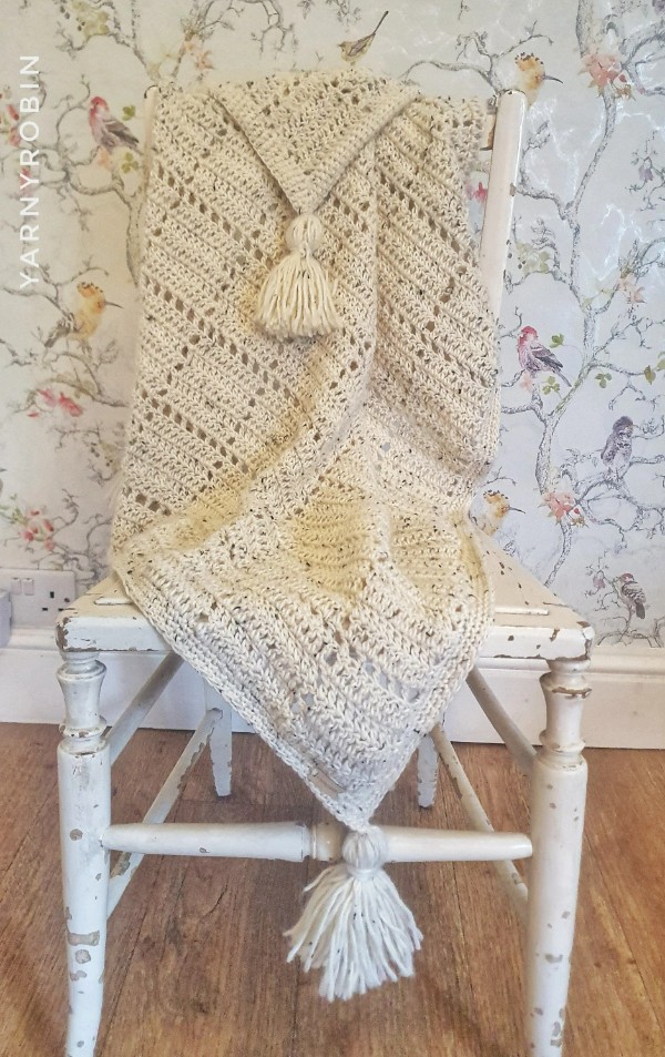 A cream coloured filet crochet blanket with chunky tassels.