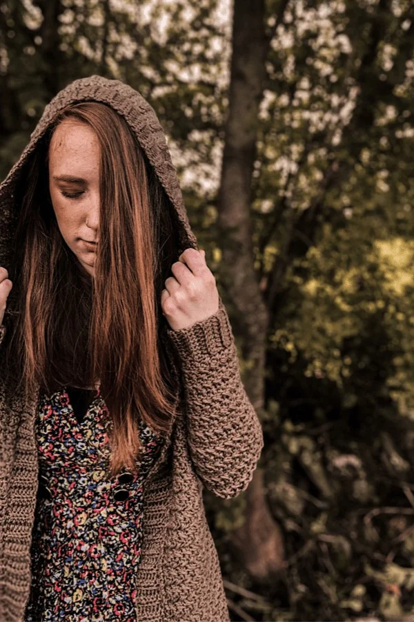 A woman wearing a brown hooded cardigan crochted with ribbed edging and cuffs.