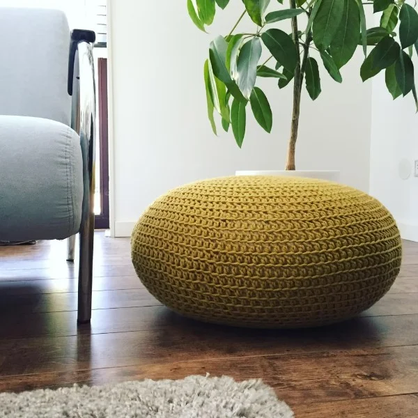 A mustard yellow crochet pouf on a timber floor with a plant in the background.