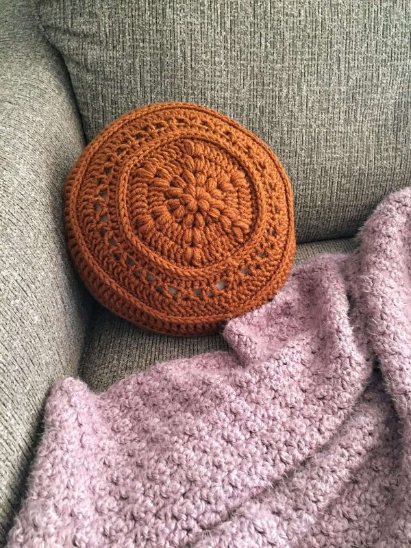 An orange coloured round crochet pillow with a floral motif.