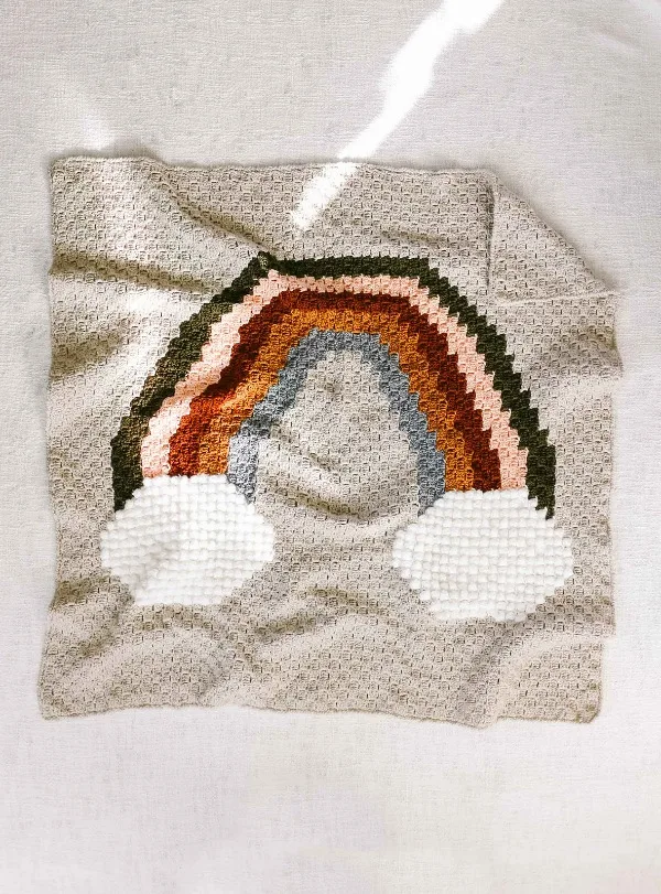 A beige c2c baby blanket with a warm toned rainbow and clouds.