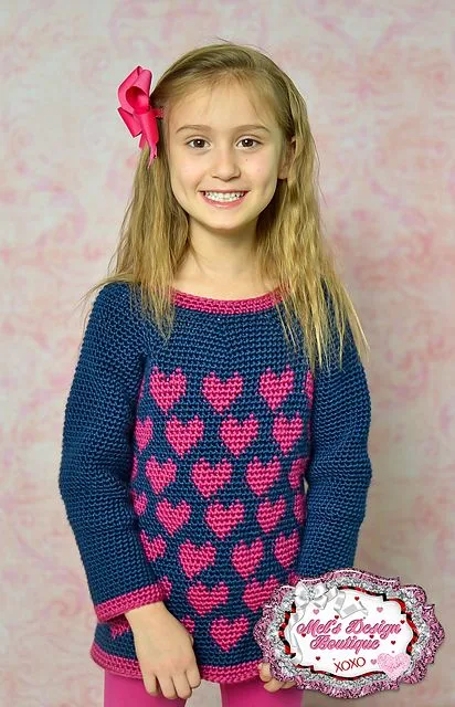 A girl wearing a navy blue crochet sweater with bright pink heart motfis.