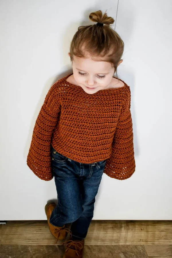 A toddler wearing a rust coloured crochet sweater with bell-shaped sleeves.