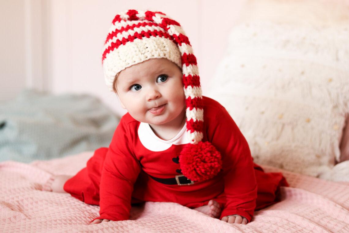 A baby in a red santa suit wearing a red and white striped crochet Christmas hat.