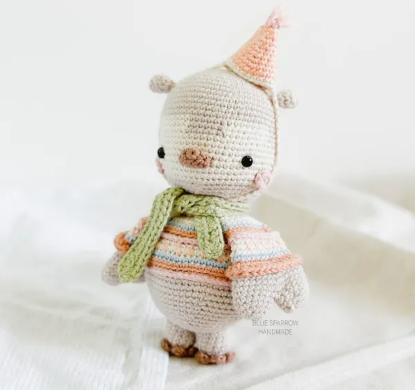 A pastel coloured crochet owl weraing a striped sweater, a scarf, and a party hat.