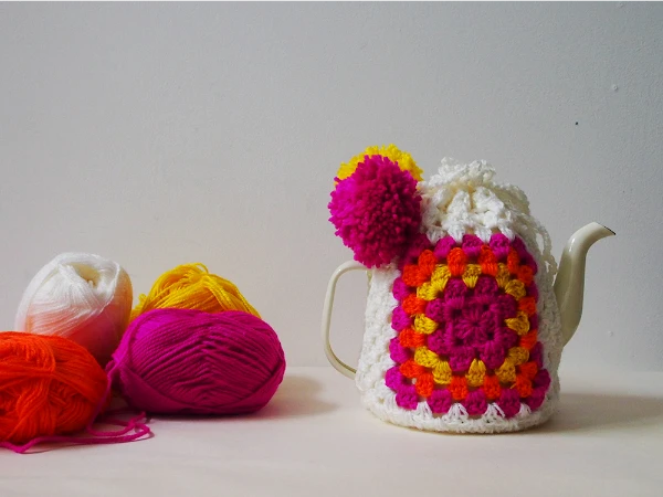 A tall crochet granny stitch teapot cover with pompom detail.