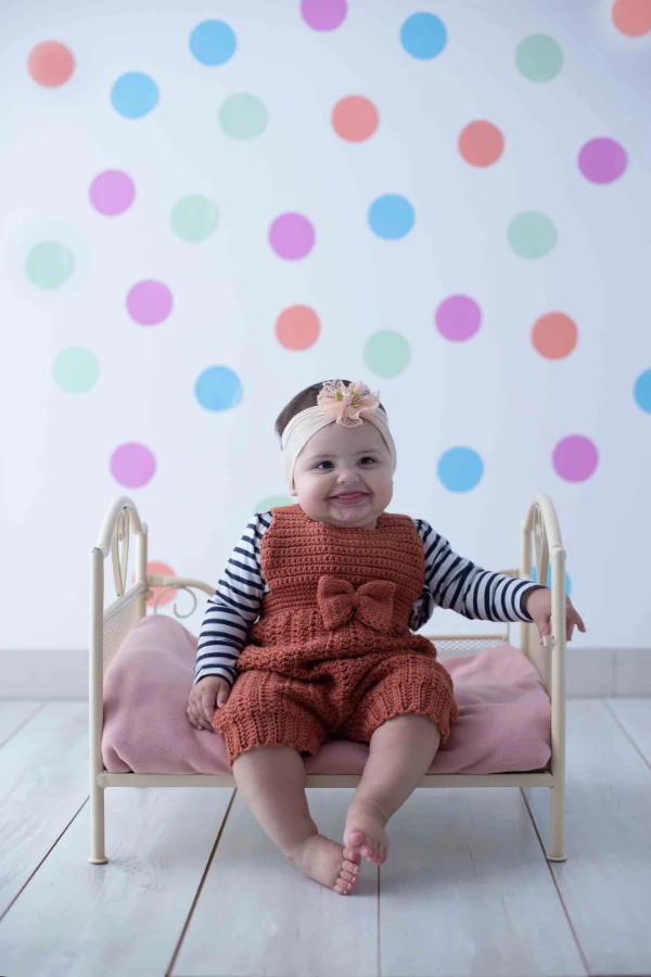 A baby wearing a striped shirt and rust coloured crochet overalls with a big bow.