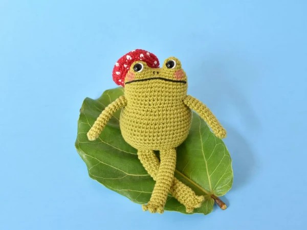A crochet frog on a leaf wearing a toadstool beret.