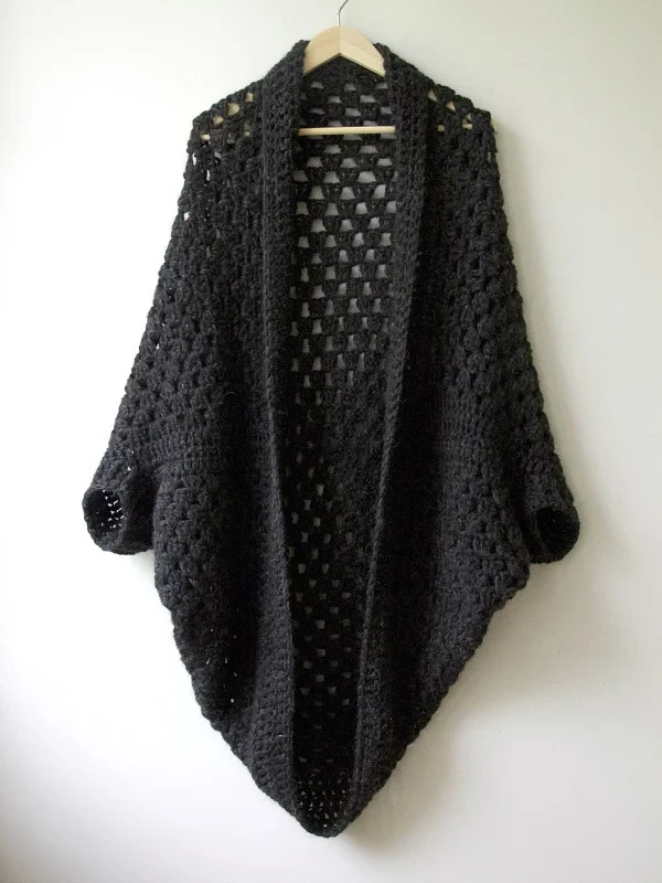 A black granny square cocoon shrug on a wooden hanger.