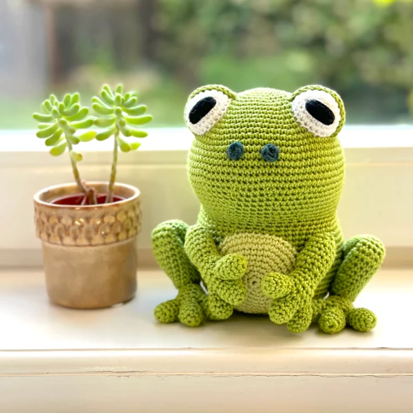 Green crochet frog amigurumi sitting next to a tiny house plant.