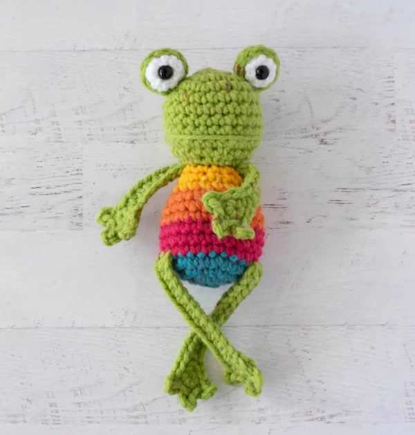 A crochet frog with a rainbow striped belly.