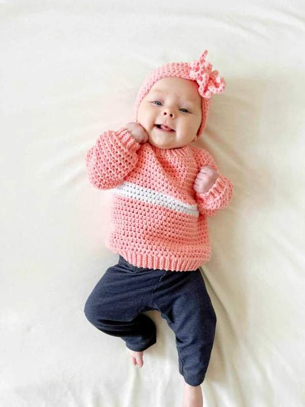 A baby girl wearing a pink crochet pullover with a white stripe.