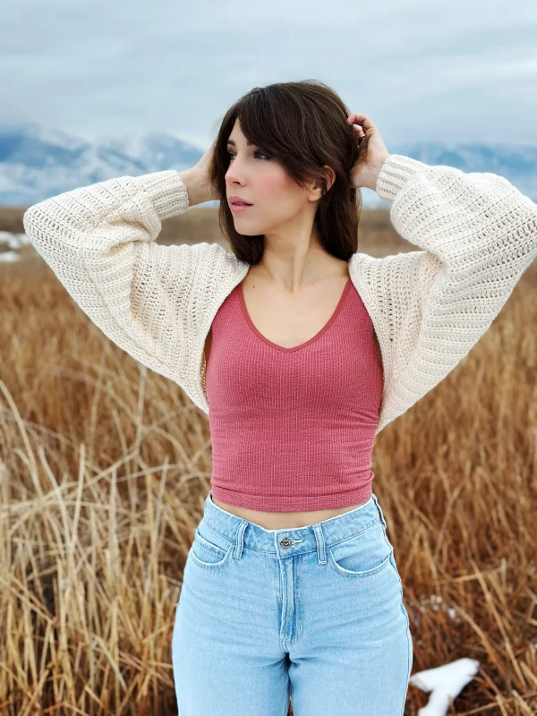 A woman in a field wearing a cream coloured crochet shrug over a pink tank.