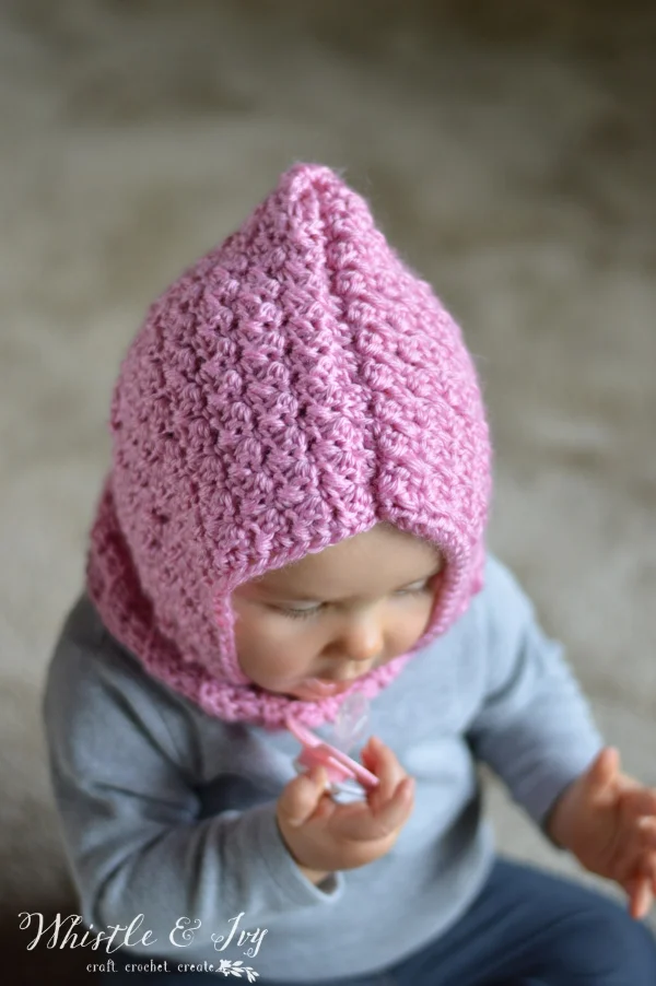 A baby wearing a pink, crocheted cowl with a hood.