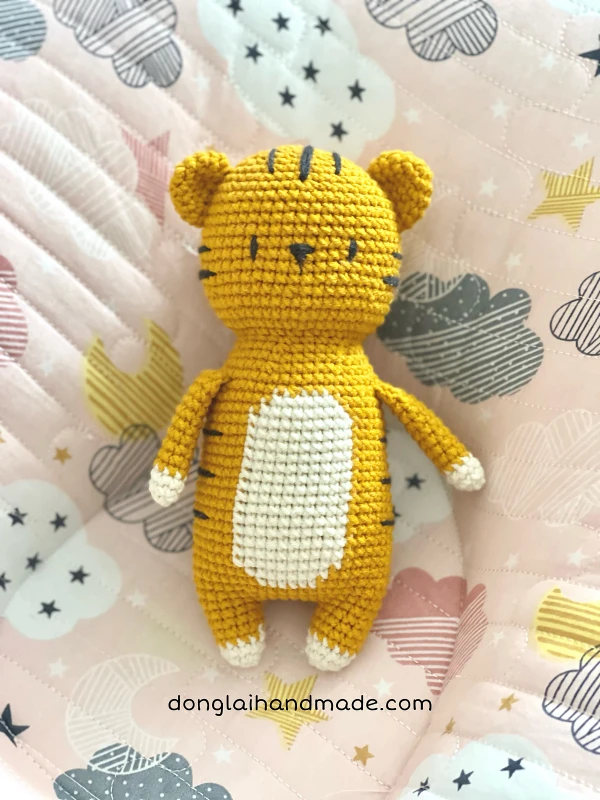 A flat lay image of a cute crochet tiger toy with embroidered facial details and stripes.