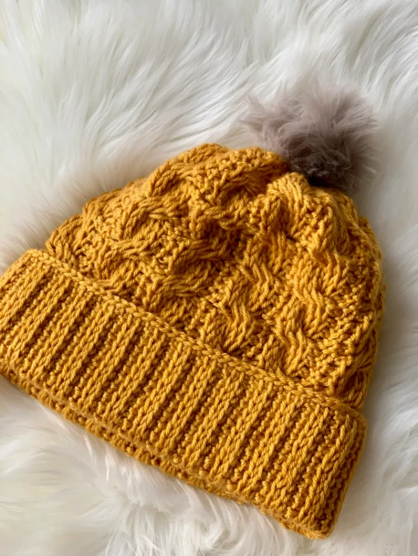 A mustard yellow Tunisian crochet hat with a faux fur pompom.