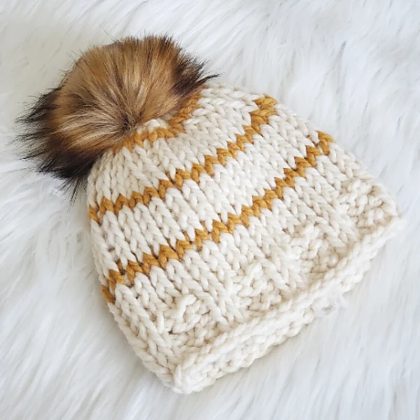 A white Tunisian Knit stitch beanie with thin yelow stripes and a faux fur pompom.