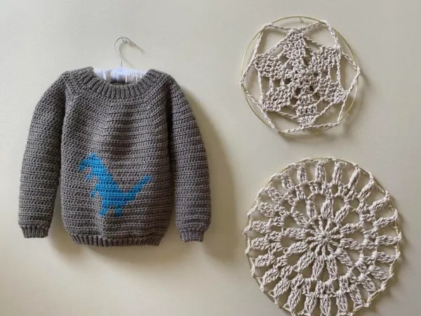 A grey childs crochet sweater with a dinosaur motif.